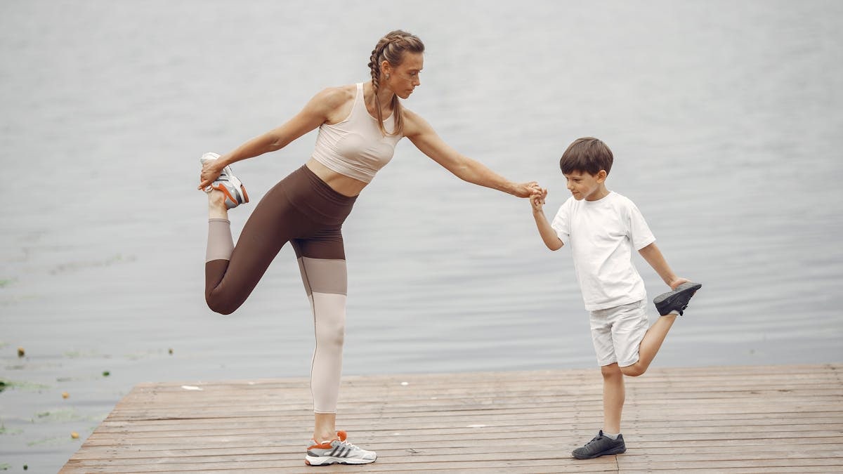 A mother and son holding a yoga pose together on a wooden pier by the water, promoting family fitness.